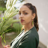 A woman with long brown hair in a ponytail, wearing a dark green top and unique jewelry—Begonia Leaves Earrings—stands outdoors in sunlight holding tall grass, looking directly at the camera. The softly blurred background is perfect for plant lovers.