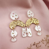 A pair of boho chic dangle earrings featuring gold textured arches and white, marble-like pieces, adorned with small colored gems, rhinestones, and displayed on soft pink fabric with dried flowers beside them.