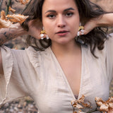 A woman with wavy brown hair poses outdoors among dry autumn leaves, wearing a light beige dress and boho chic dangle earrings adorned with rhinestones, her hands touching her hair and an intense, confident expression on her face.
