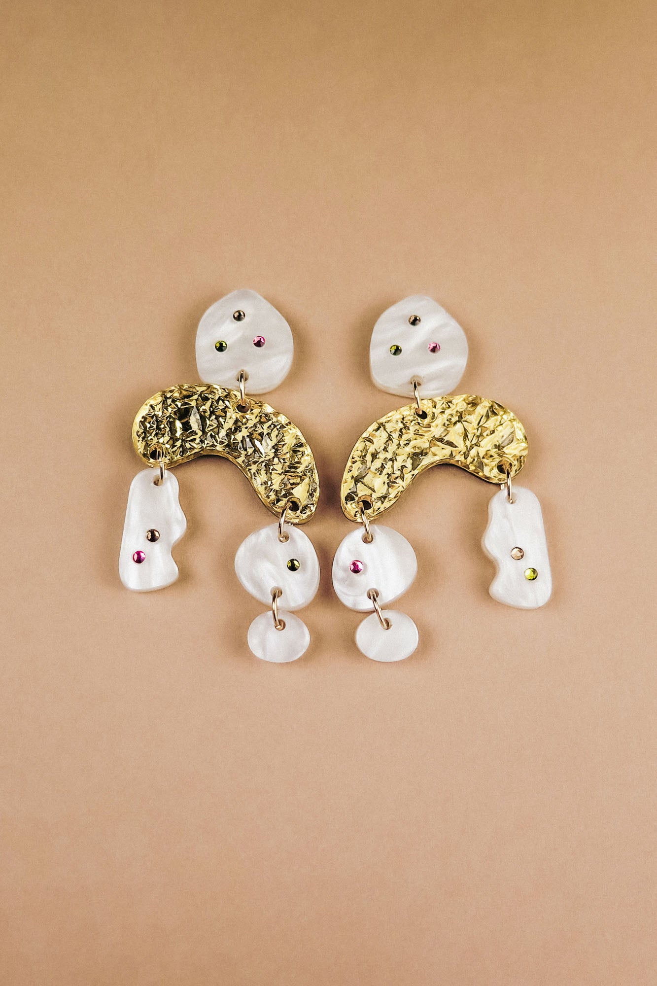 A pair of abstract dangle earrings with textured gold curved pieces, irregular white shapes, and small embedded red and green rhinestones, displayed on a beige background.