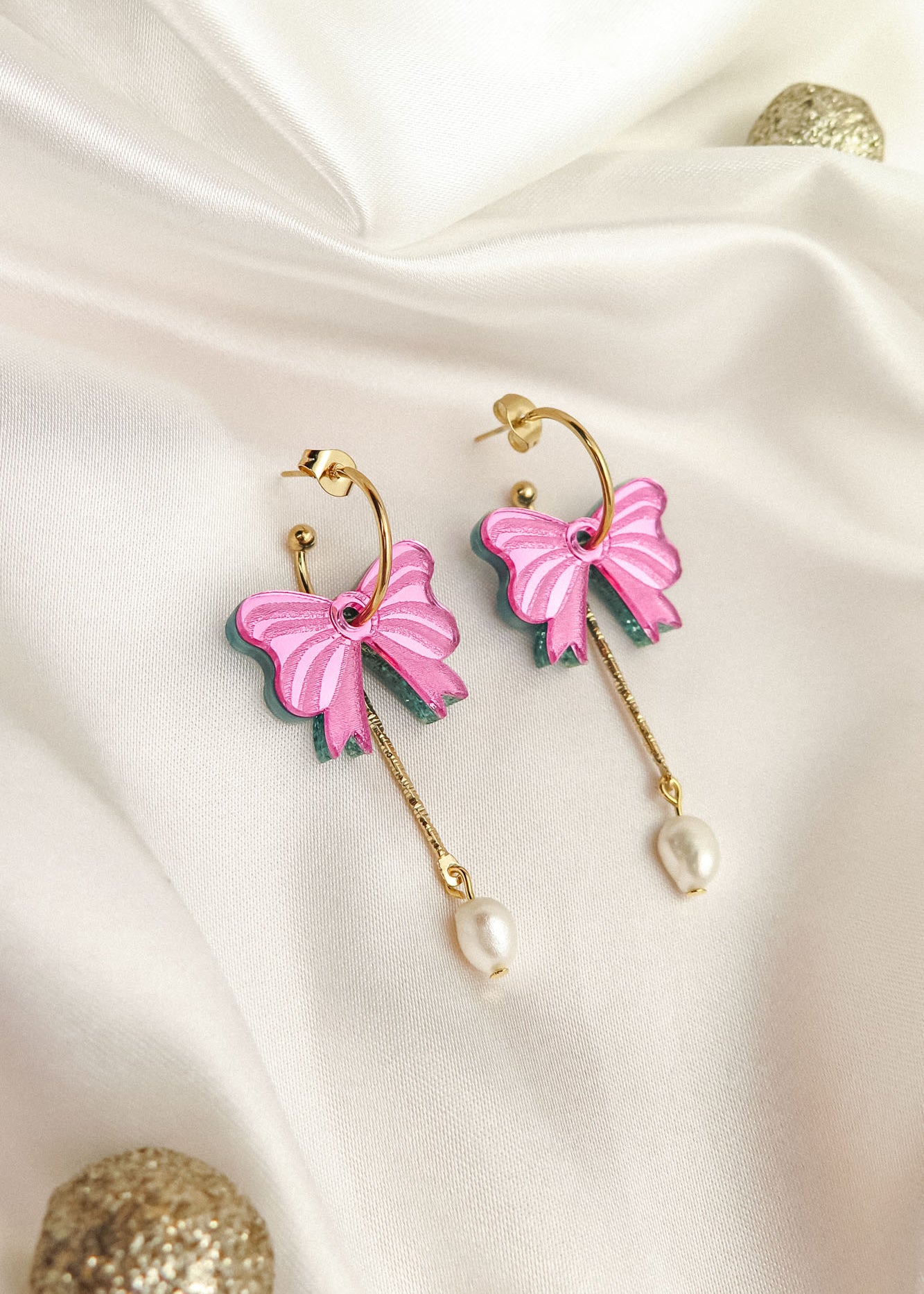 A pair of gold-plated c-hoop earrings with pink butterfly charms and freshwater pearl drops, displayed on white satin fabric with decorative gold spheres nearby.