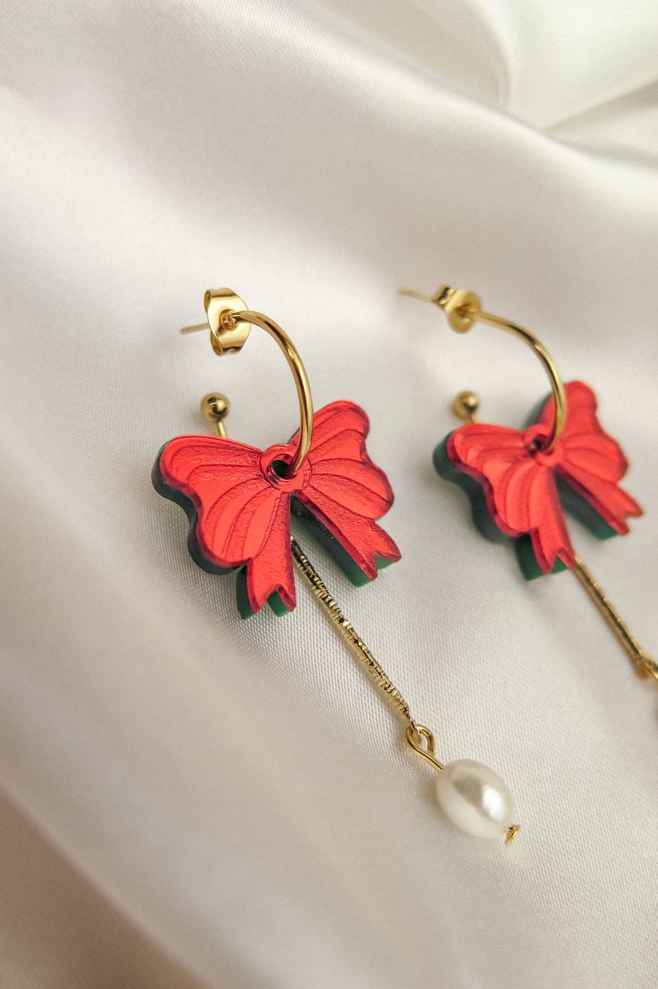 A pair of gold-plated c-hoop earrings with red butterfly-shaped charms and dangling gold chains ending in a single freshwater pearl, displayed on white satin fabric.