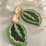 A pair of Calathea Medallion Earrings shaped like green leaves with gold edges and a touch of purple in the center, displayed on a light fabric background with delicate white foliage—nature-inspired jewelry at its finest.