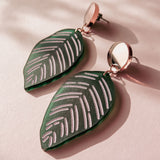 A pair of green, leaf-shaped handcrafted earrings with silver detailing and round metallic studs, inspired by Calathea Ornata Earrings, displayed on a pale pink surface with soft shadows.