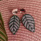 A pair of handcrafted earrings, the Calathea Ornata Earrings, with metallic round studs and dark green leaf-shaped pendants featuring light pink vein patterns, are displayed on a textured pink woven surface with green leaves in the background—a true piece of tropical nature jewelry.