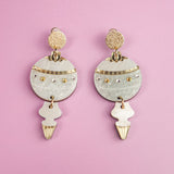 A pair of ornate, gold and white festive earrings with circular and abstract shapes, resting on a pastel purple background.
