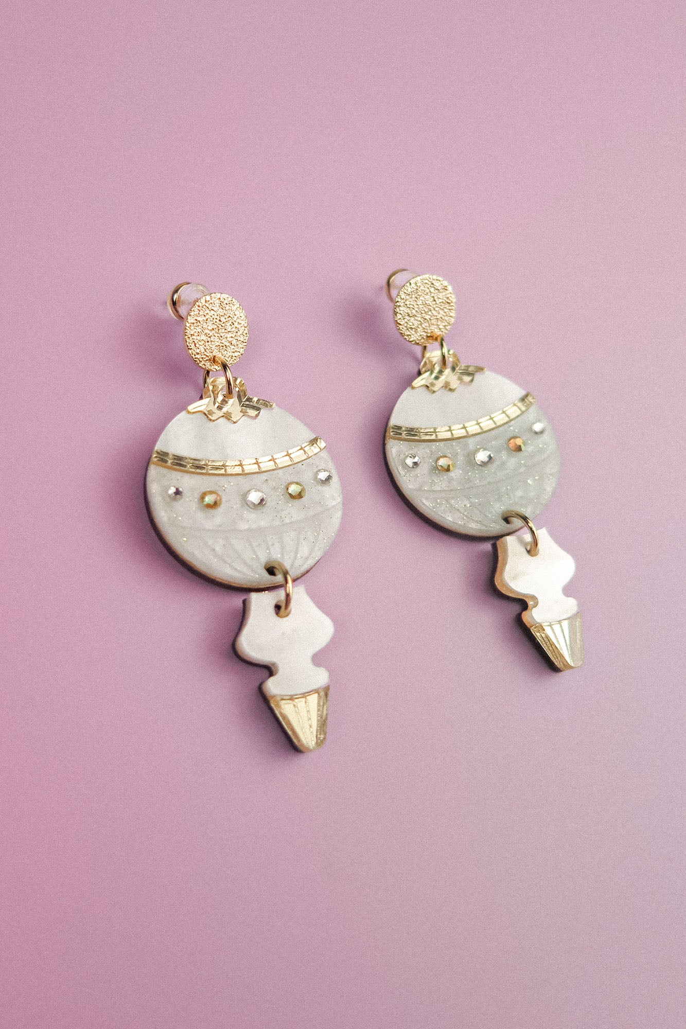 A pair of decorative dangle earrings with gold and silver accents, featuring round and arrow-shaped elements, make for stunning festive earrings, beautifully displayed on a light purple background.