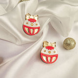 A pair of round enamel pins with red, white, and gold details, resembling festive ornaments with bows, are displayed on white satin fabric with gold glittery balls—perfect as holiday jewelry or a festive earrings-inspired accent.