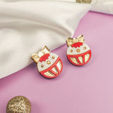 Two round festive earrings with red, white, and gold details shaped like ornaments with bows are displayed on a light purple surface next to a white cloth and glittery decorative balls—perfect holiday jewelry for any occasion.