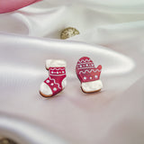 Red and white decorated cookies shaped like a Christmas stocking and mitten sit on silky white fabric, surrounded by festive accessories—a pink ribbon, gold ornaments, and charming Christmas Knitted Studs holiday earrings in the background.