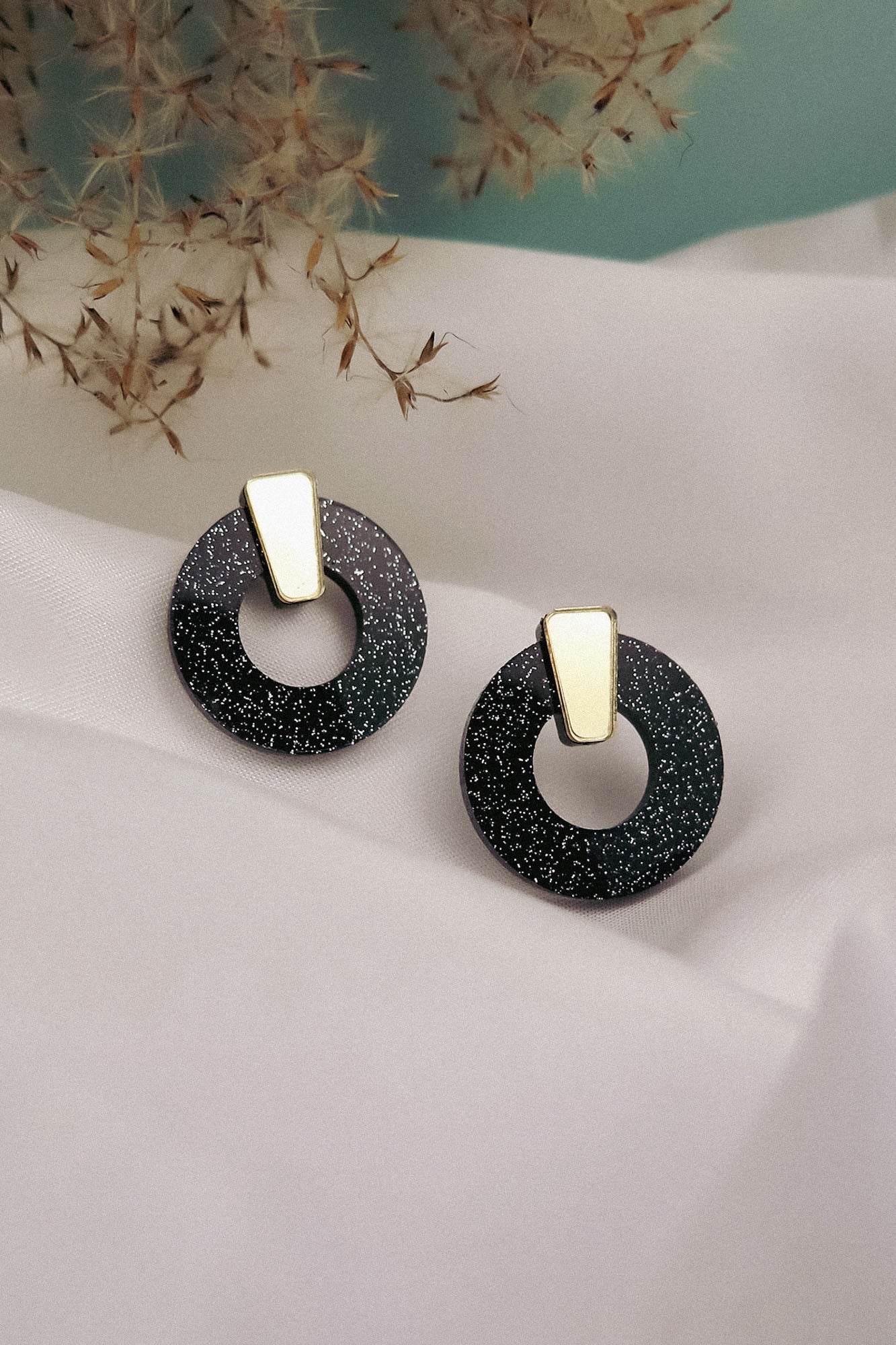 A pair of minimalist circle stud earrings in black acrylic glass with white speckles and gold triangular studs, displayed on white fabric with dried grasses in the background. Hypoallergenic for comfortable, everyday wear.