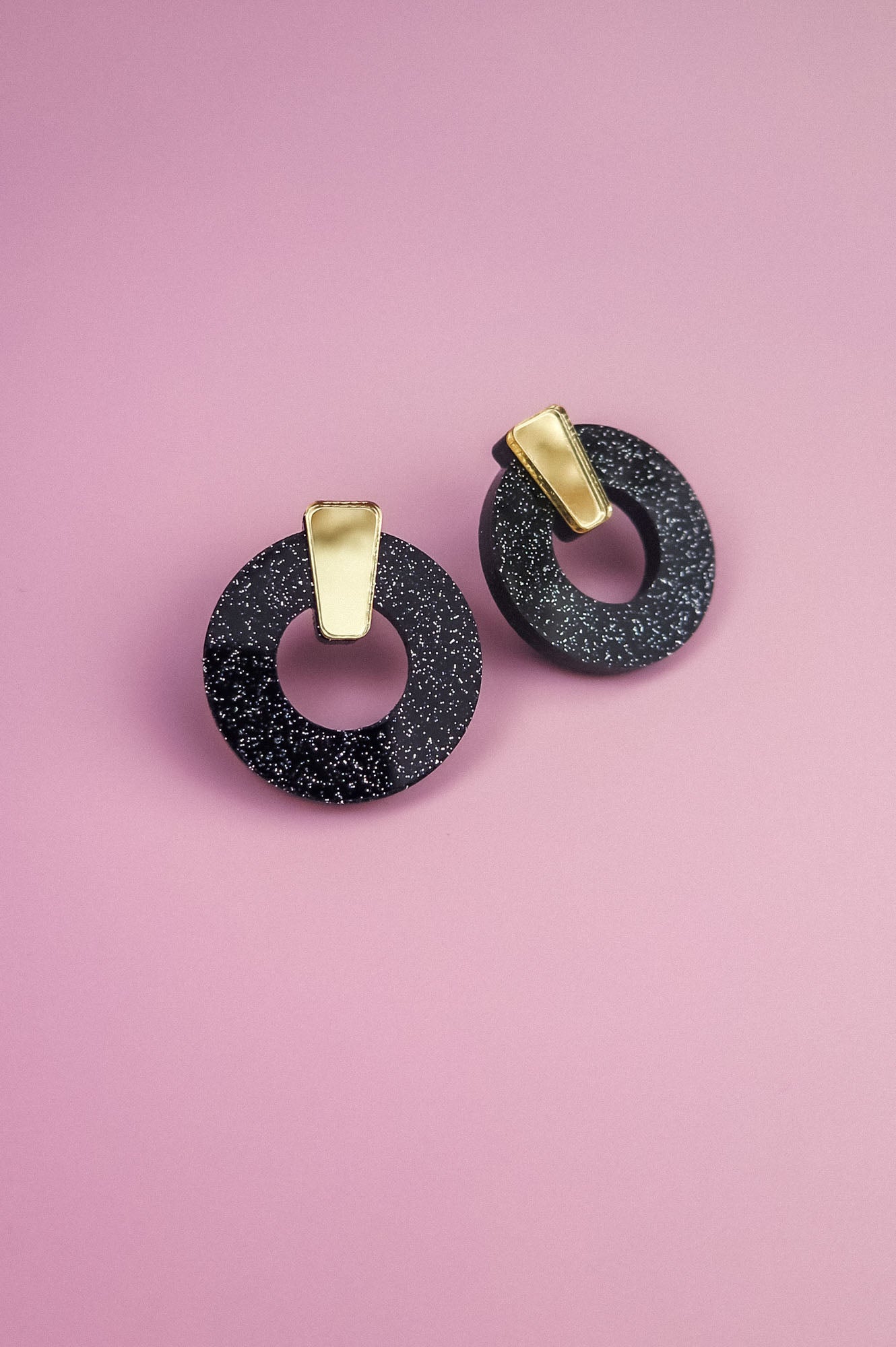A pair of minimalist circle stud earrings, crafted from hypoallergenic acrylic glass with white speckles and gold rectangular accents, are displayed on a pink background.