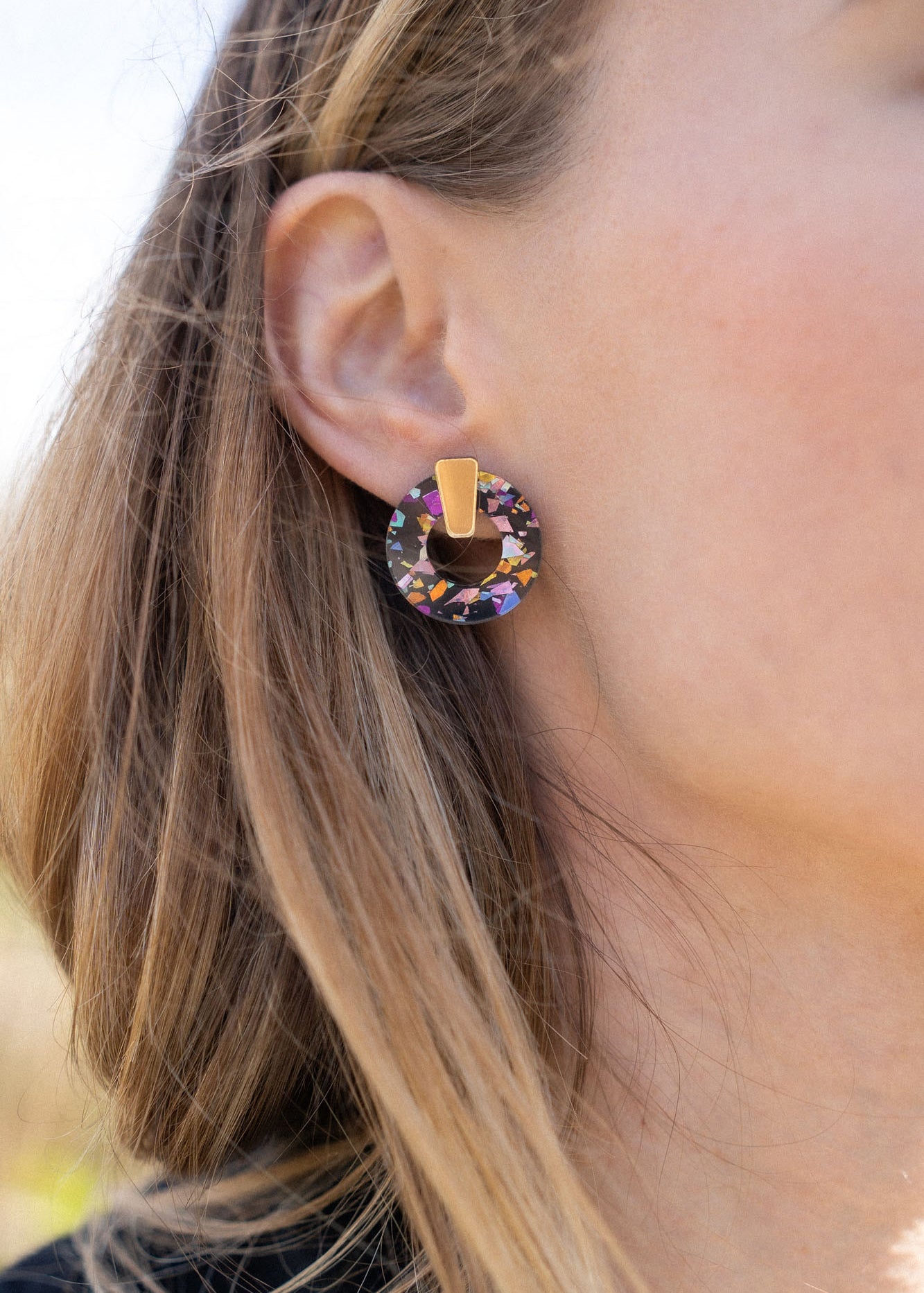 A close-up of a woman wearing Minimalist Circle Stud Earrings crafted from hypoallergenic materials, featuring a large, circular, multicolored gemstone with a gold accent, shown from the side against a blurred outdoor background.