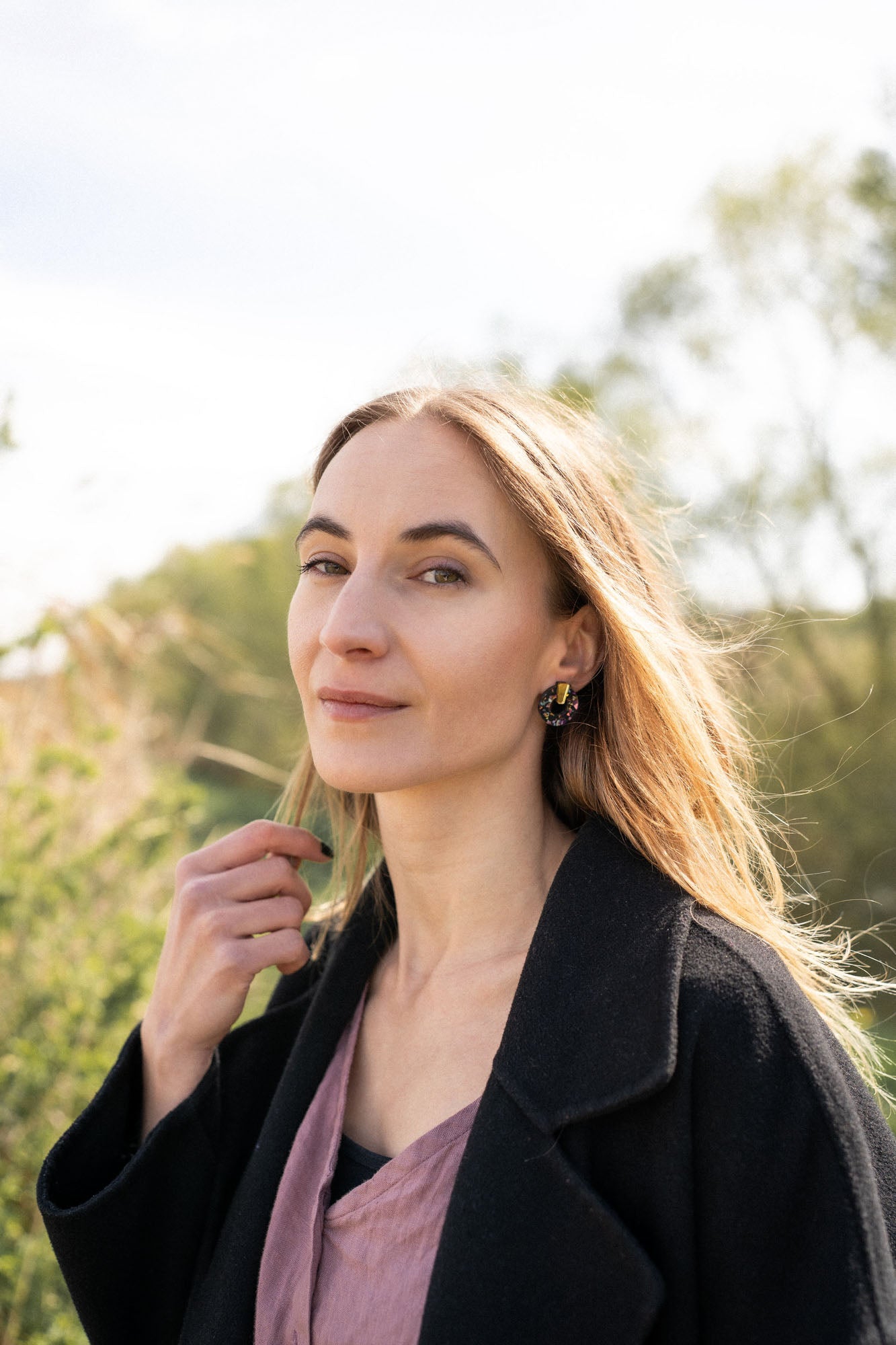 A woman with long, light brown hair stands outdoors, wearing a dark coat, mauve top, and minimalist circle stud earrings. She lightly touches her hair and looks calmly at the camera against green foliage and a bright, sunlit sky.