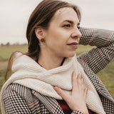 A woman in a houndstooth coat and cream scarf, embodying minimalist elegance, stands outdoors on a grassy, overcast day. She gazes thoughtfully into the distance, one hand on her chest and the other behind her head.