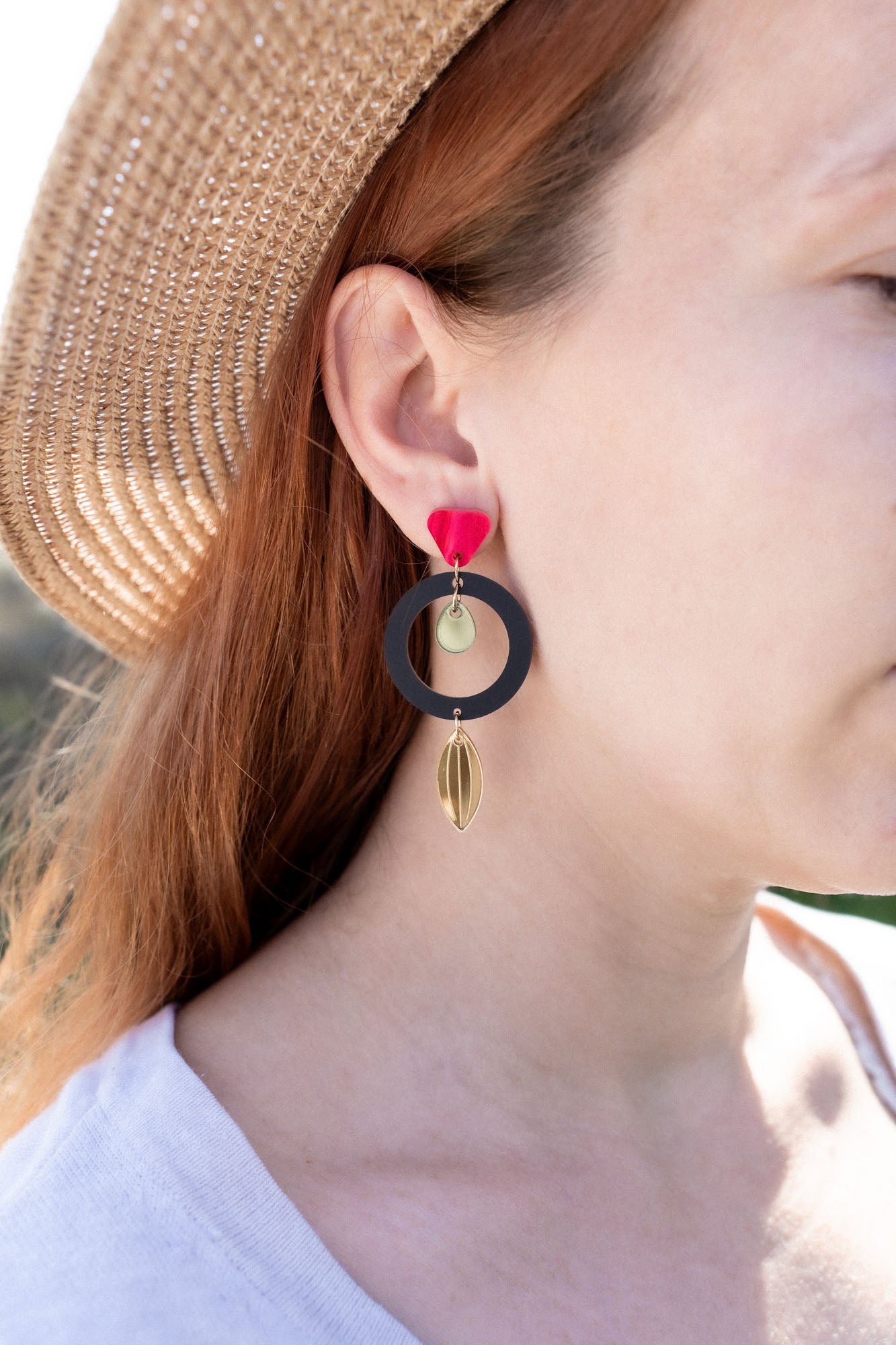 A woman in a straw hat and white shirt showcases artisan jewelry: dangle earrings crafted from recycled acrylic, featuring a black circle, gold shapes, and a red accent near the ear for a striking statement look.