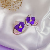 Crocus Stud Earrings shaped like hand-painted flowers with purple petals and yellow centers, displayed on white satin fabric with dried grasses in the background. These beautiful earrings are allergy-free.