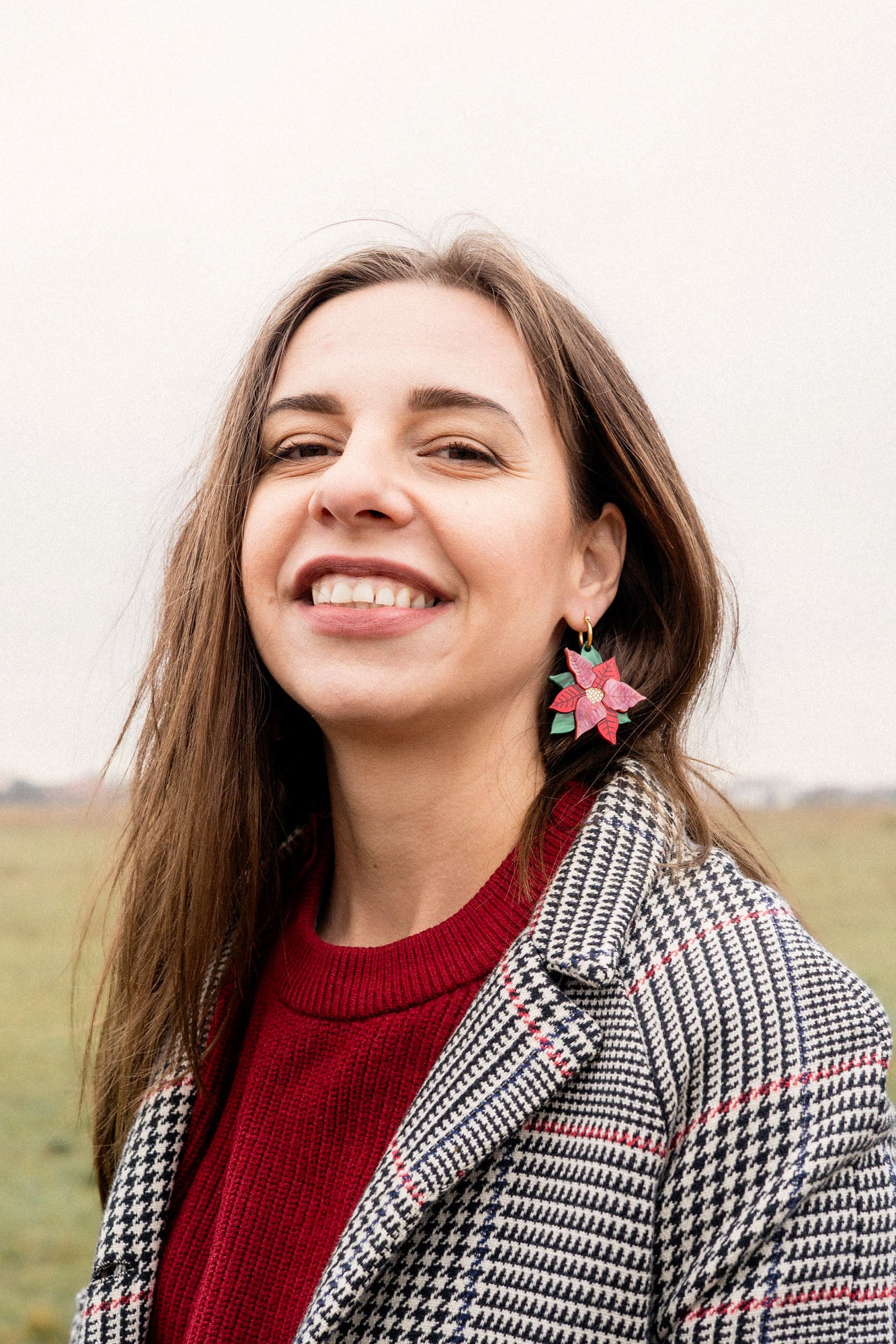 A woman with long brown hair smiles outdoors, wearing a red sweater, a black-and-white checkered coat, and festive holiday earrings. The background is a grassy field under an overcast sky.