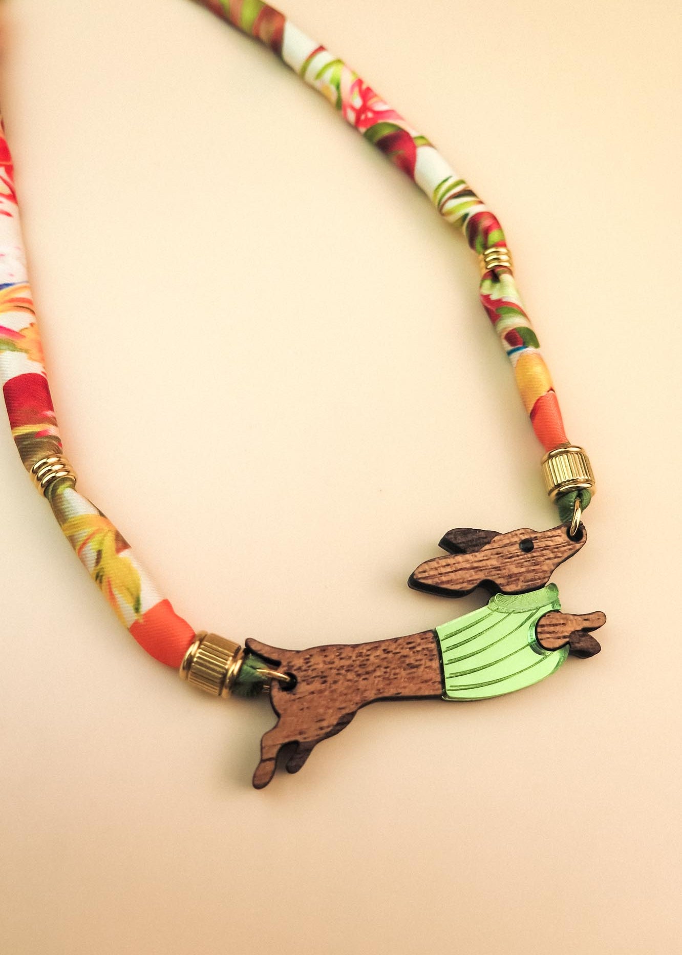 A piece of handcrafted jewelry featuring a colorful, patterned fabric cord and a wooden pendant shaped like a leaping rabbit in a green shirt. Made with recycled materials, set against a soft beige background.