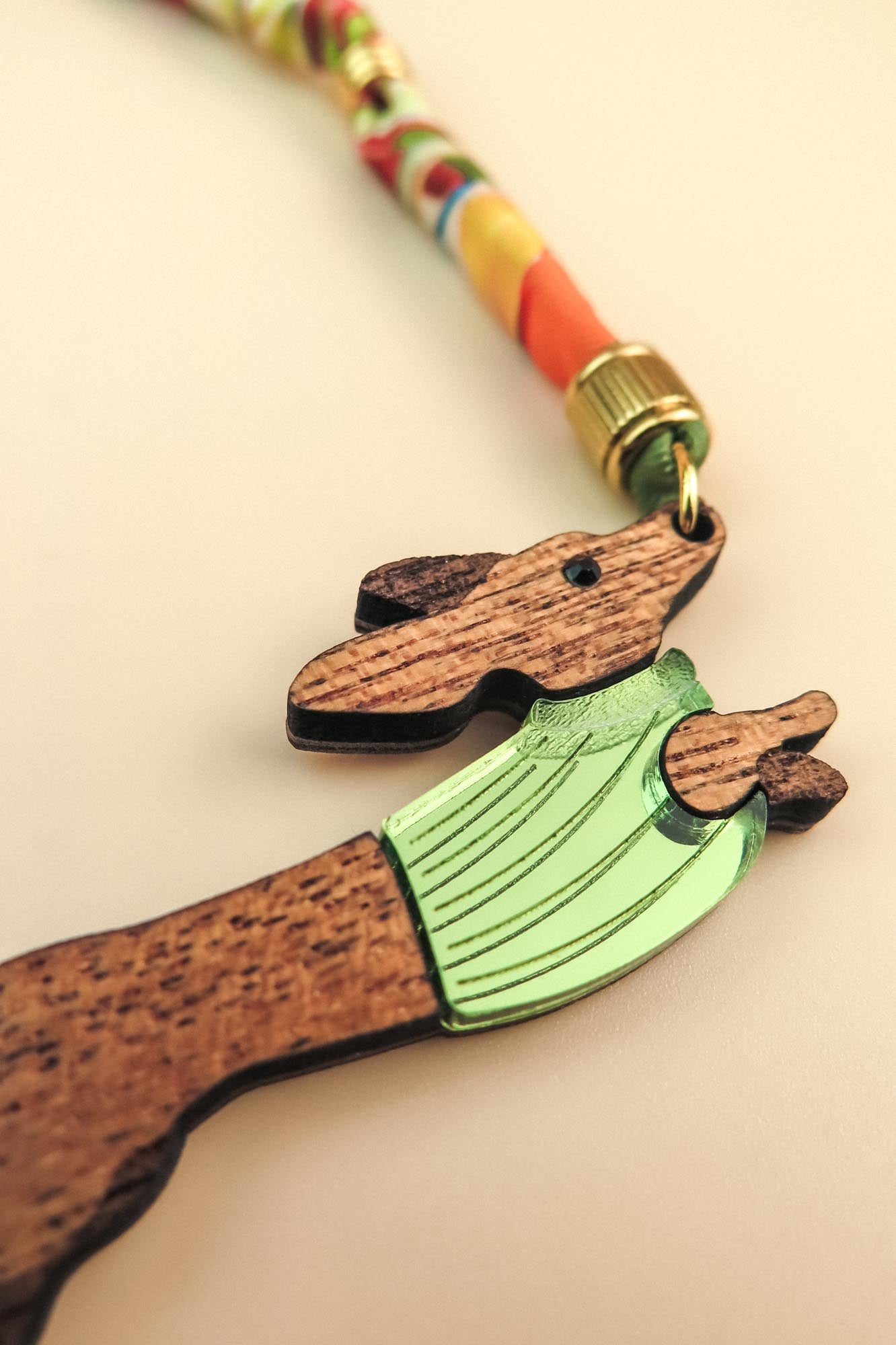 A close-up of a handcrafted wooden keychain shaped like a dog in a green sweater, attached to a colorful, twisted cord with a gold-colored metal clasp.