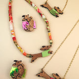 Colorful handcrafted jewelry featuring Dachshund necklaces and brooches with dogs in green and pink sweaters, set against a pale beige background. Each piece is thoughtfully made using recycled materials.