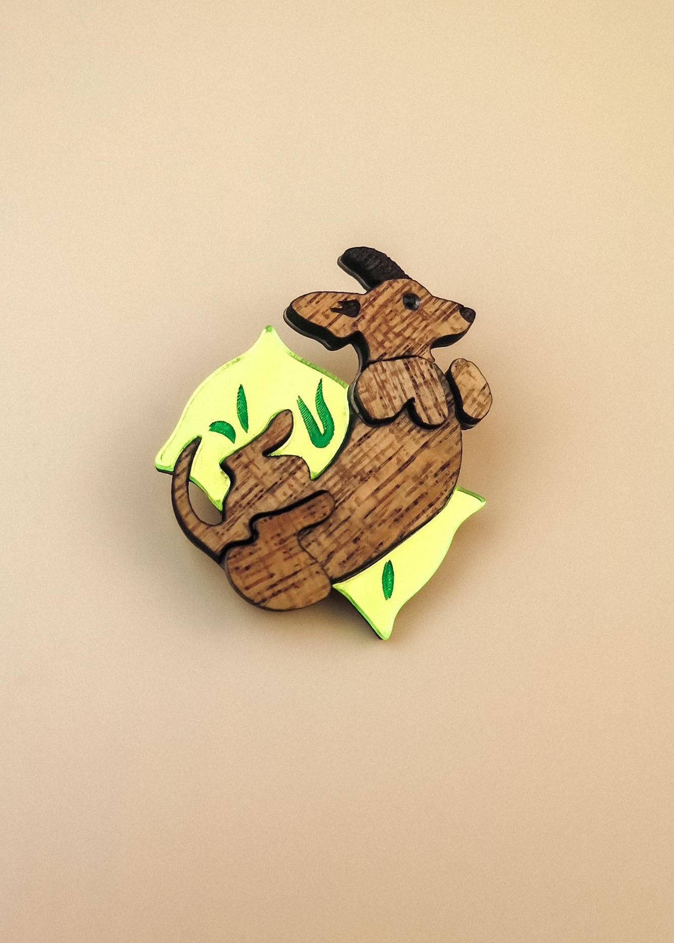 A playful brooch shaped like a kangaroo lies on a plain beige background. With engraved details and bright yellow and green leaf-like accents, this wooden piece adds whimsy to any dog accessory collection.