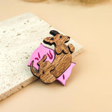 A playful Dachshund brooch shaped like a curled-up dog holding a pink, leaf-shaped object rests on a textured white stone slab against a soft beige background.