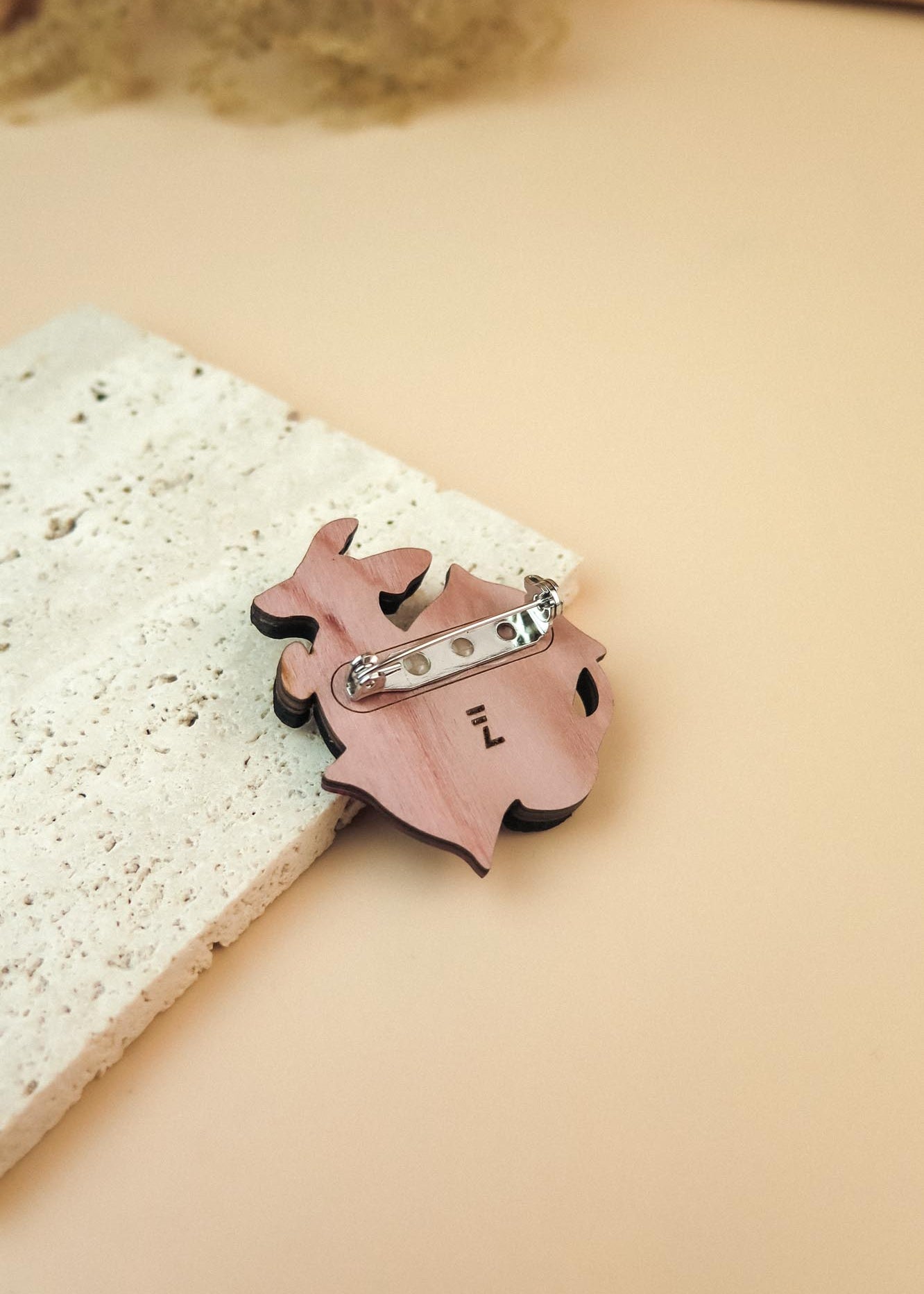 A pink, animal-shaped dog pin with a silver clasp lies on a textured, off-white stone surface against a soft beige background—perfect for lovers of playful jewelry.