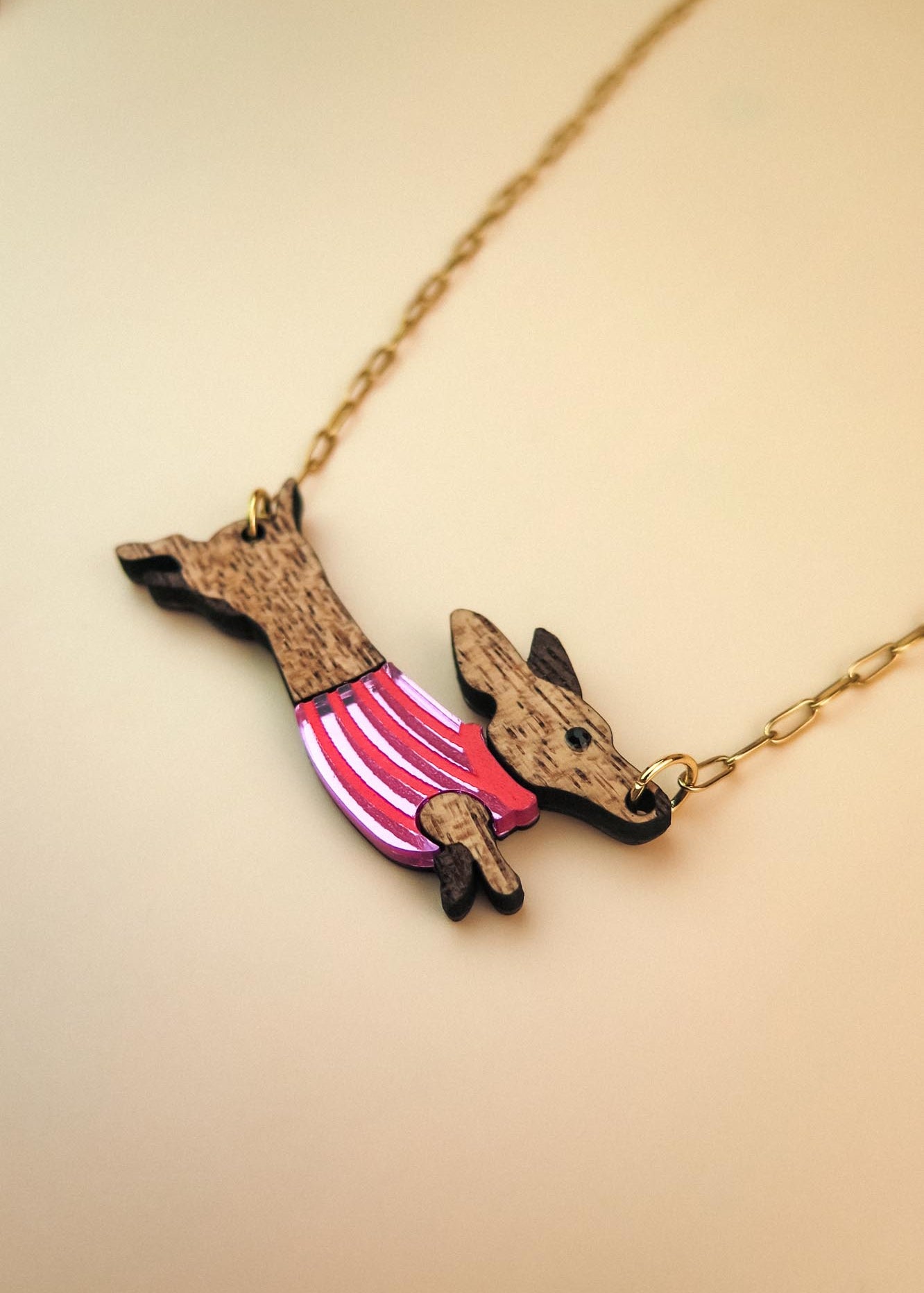 A necklace with a gold chain featuring a pendant shaped like a Dachshund in a colorful sweater, crafted from sustainable wood, set against a light beige background.