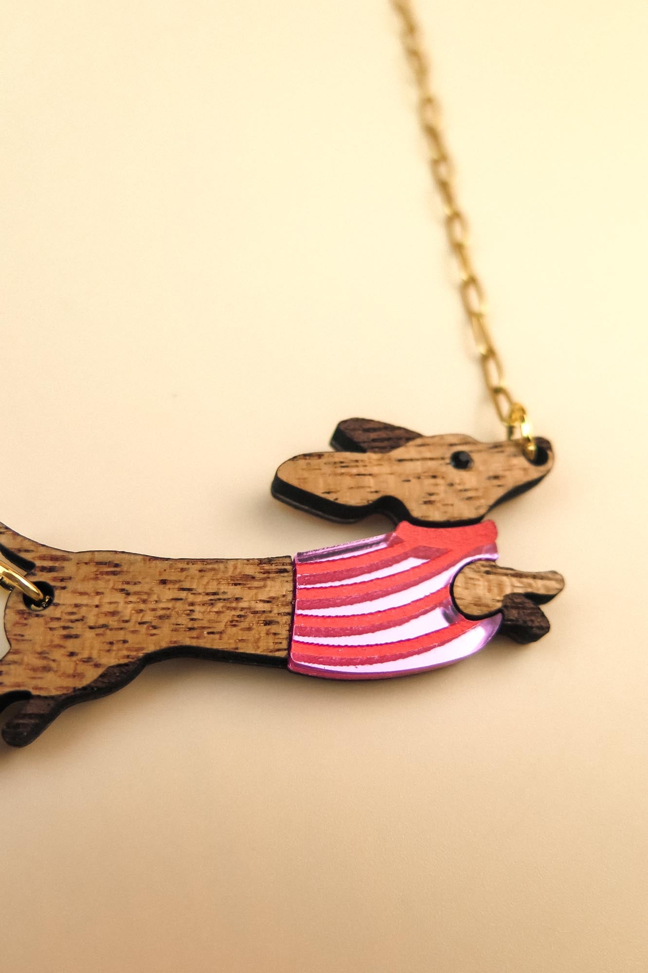 A wooden necklace pendant shaped like a dachshund dog in a colorful sweater, crafted from sustainable wood and attached to a gold chain, shown on a light beige background.