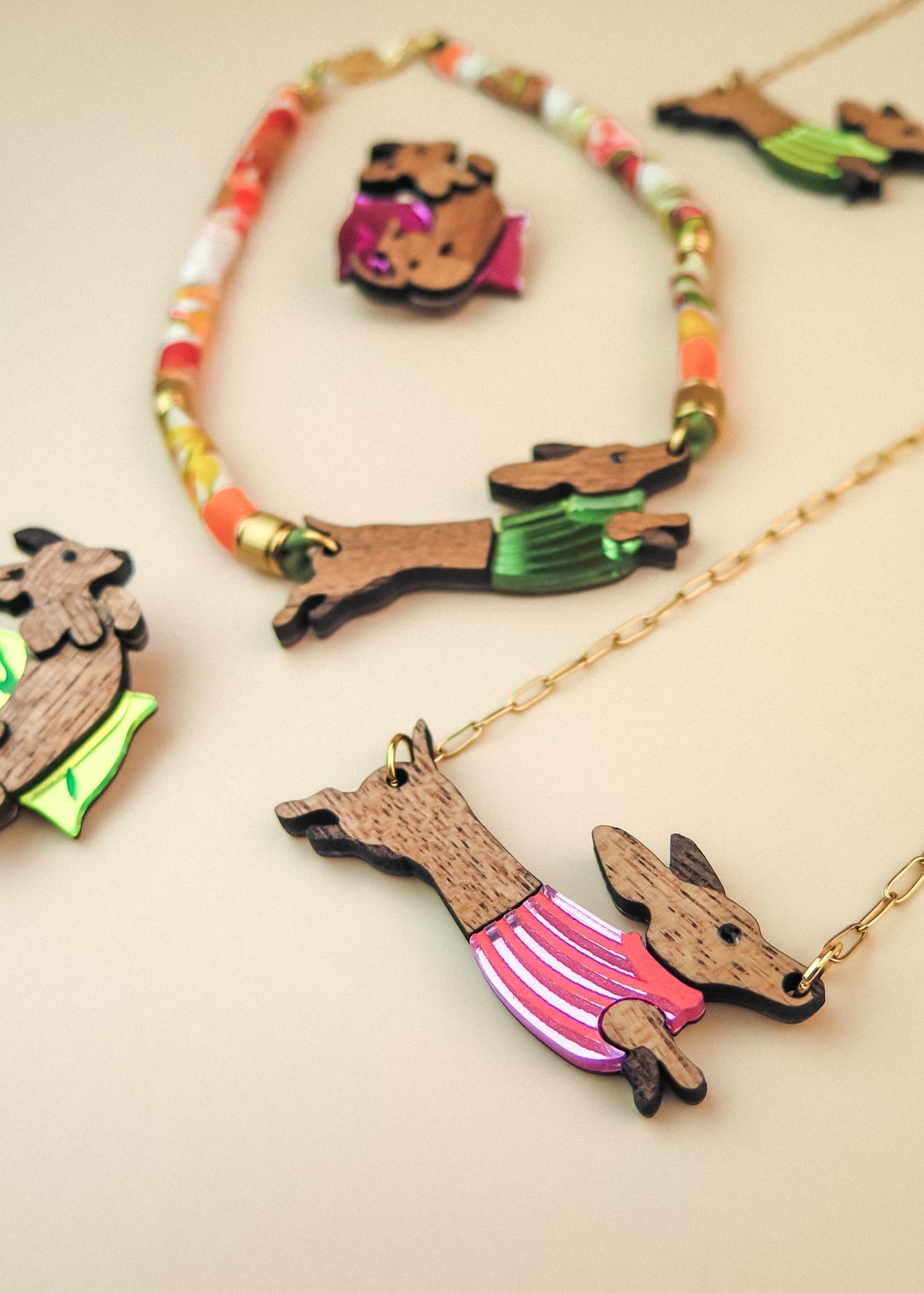 Colorful jewelry featuring Dachshund-shaped pendants crafted from sustainable wood, each dressed in a bright, colorful sweater. Displayed on a beige surface, the collection includes playful necklaces and pins with gold chains and vibrant beads.