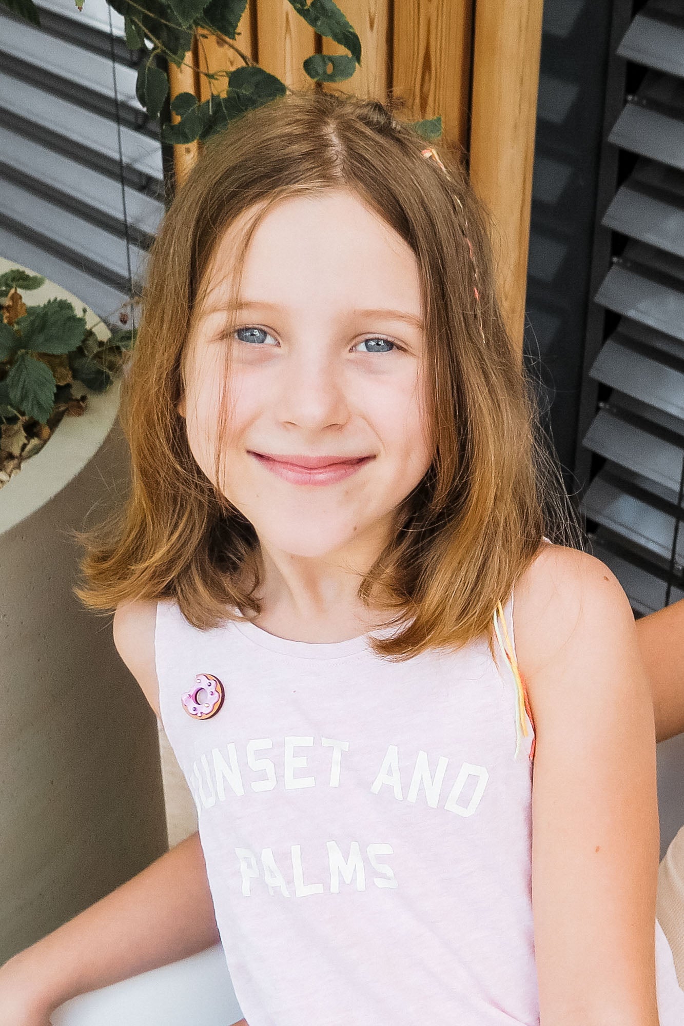 A young girl with shoulder-length light brown hair and blue eyes smiles at the camera. She wears a pale pink sleeveless top reading SUNSET AND PALMS, paired with a handcrafted Donut Pin. Plants and wooden details decorate the background.