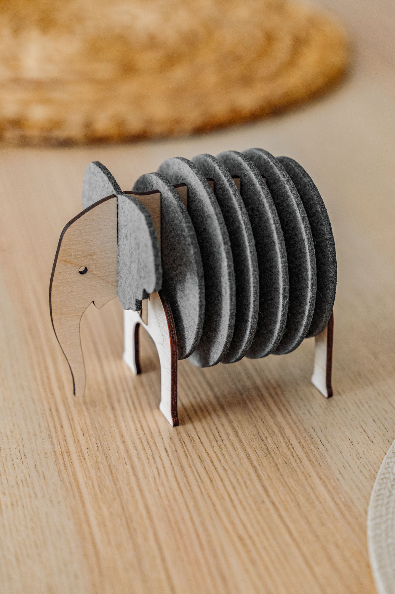 A set of round, gray Elephant Coasters is stacked vertically in a wooden holder shaped like an elephant, adding witty design and furniture protection to your light wood table.