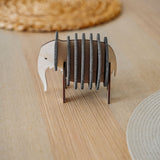 A small, elephant-shaped coaster holder made of wood and dark felt stands on a light wooden table, showcasing witty design for furniture protection, surrounded by round woven placemats.
