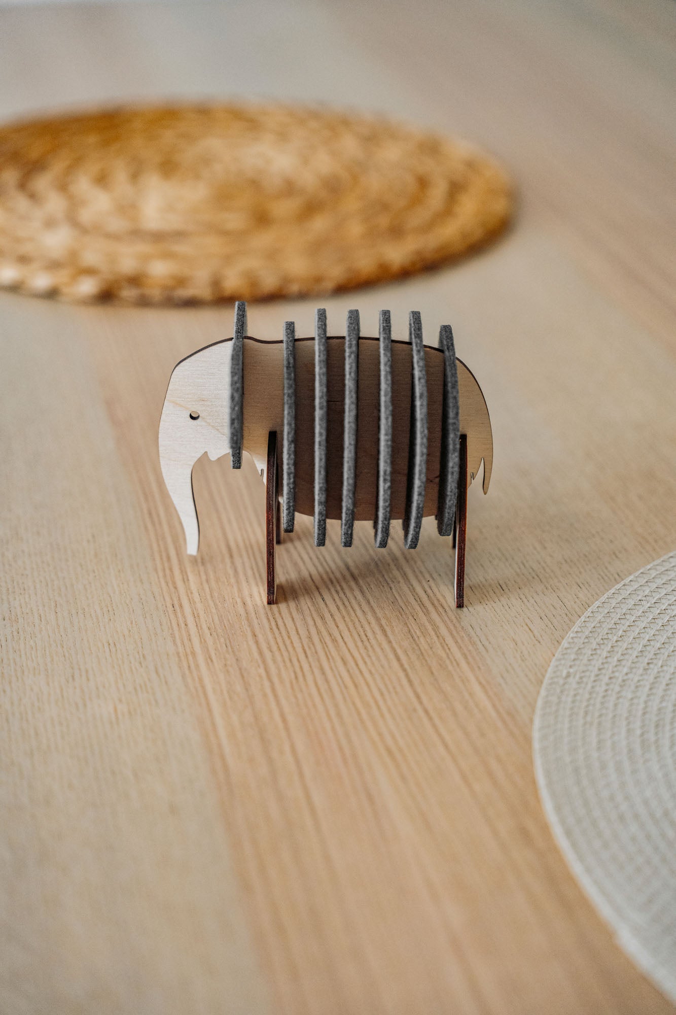 A small, elephant-shaped coaster holder made of wood and dark felt stands on a light wooden table, showcasing witty design for furniture protection, surrounded by round woven placemats.