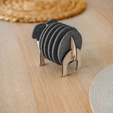 A set of round black Elephant Coasters is neatly arranged in a witty design sheep-shaped wooden holder, placed on a light wooden table with woven placemats in the background for stylish furniture protection.