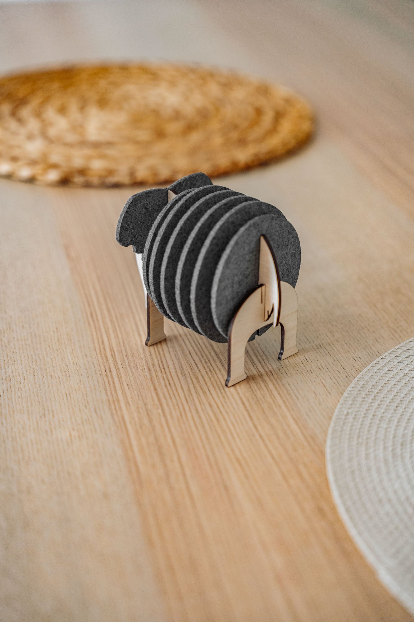 A set of round black Elephant Coasters is neatly arranged in a witty design sheep-shaped wooden holder, placed on a light wooden table with woven placemats in the background for stylish furniture protection.