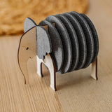 A wooden and felt Elephant Coasters holder with witty design stands on a wooden table, offering both charm and furniture protection, while a round woven placemat is blurred in the background.