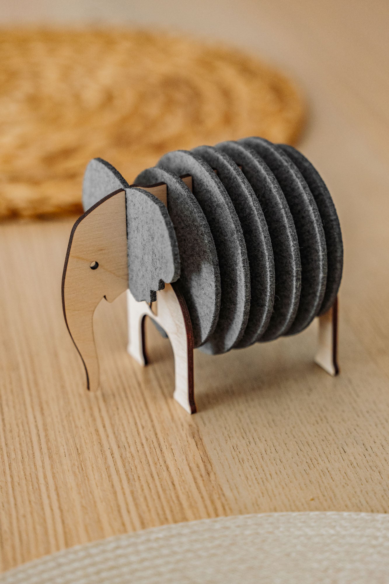 A wooden and felt Elephant Coasters holder with witty design stands on a wooden table, offering both charm and furniture protection, while a round woven placemat is blurred in the background.