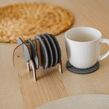 A set of round Elephant Coasters with witty design sits in a wooden elephant-shaped holder on a table, offering furniture protection while a white mug rests on one coaster.