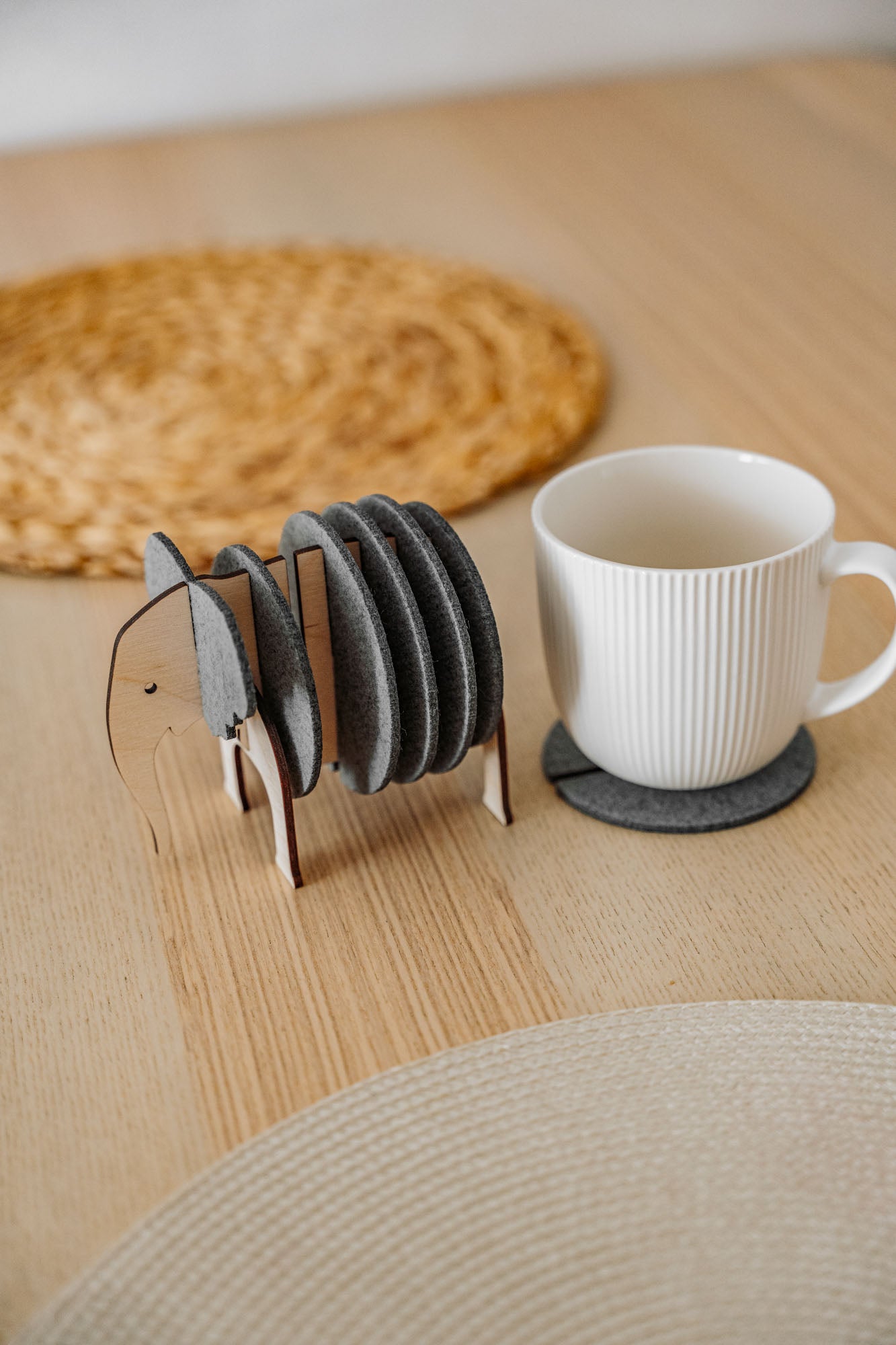 A set of round Elephant Coasters with witty design sits in a wooden elephant-shaped holder on a table, offering furniture protection while a white mug rests on one coaster.