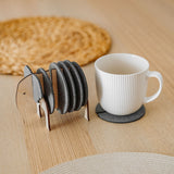 A white ribbed coffee cup sits on a round gray coaster. Beside it, Elephant Coasters with witty design are stacked in a wooden elephant-shaped holder, providing both style and furniture protection on the light wooden table. A woven placemat is in the background.