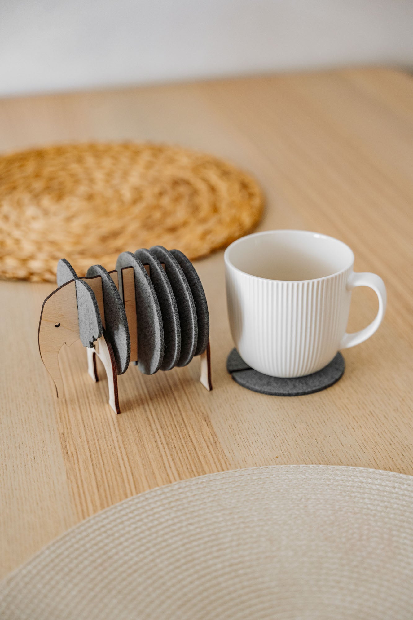 A white ribbed coffee cup sits on a round gray coaster. Beside it, Elephant Coasters with witty design are stacked in a wooden elephant-shaped holder, providing both style and furniture protection on the light wooden table. A woven placemat is in the background.