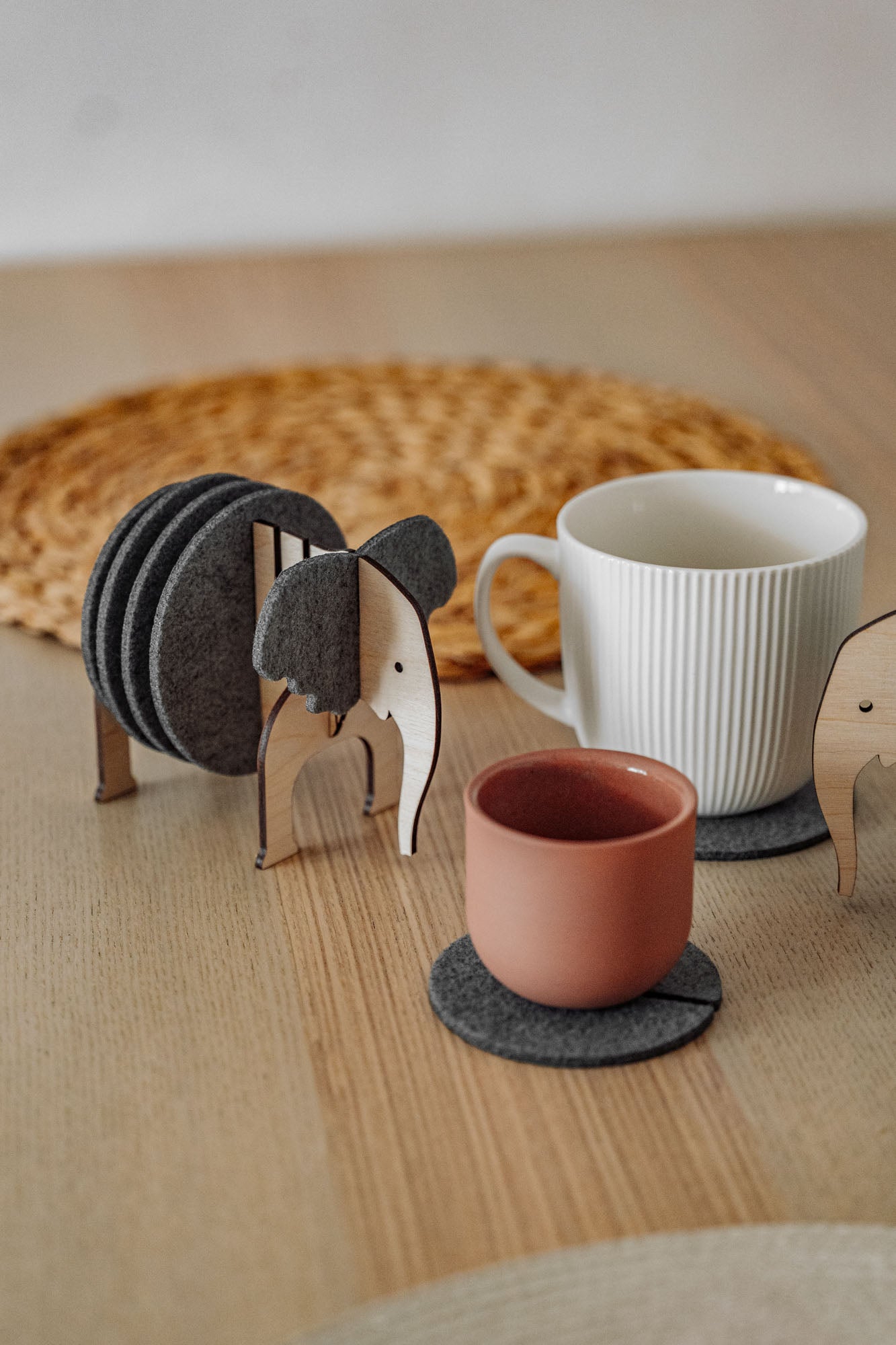 A set of Elephant Coasters with witty design offers stylish furniture protection, nestled in a wooden elephant-shaped holder on a wooden table beside a pink ceramic cup, white mug, and woven placemat.