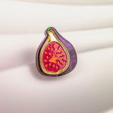 A colorful Fig Pin shaped like a fig, with a purple outer edge and a vibrant red and yellow interior, is displayed on a softly lit, white, textured background—a handcrafted, lightweight accessory perfect for any collection.