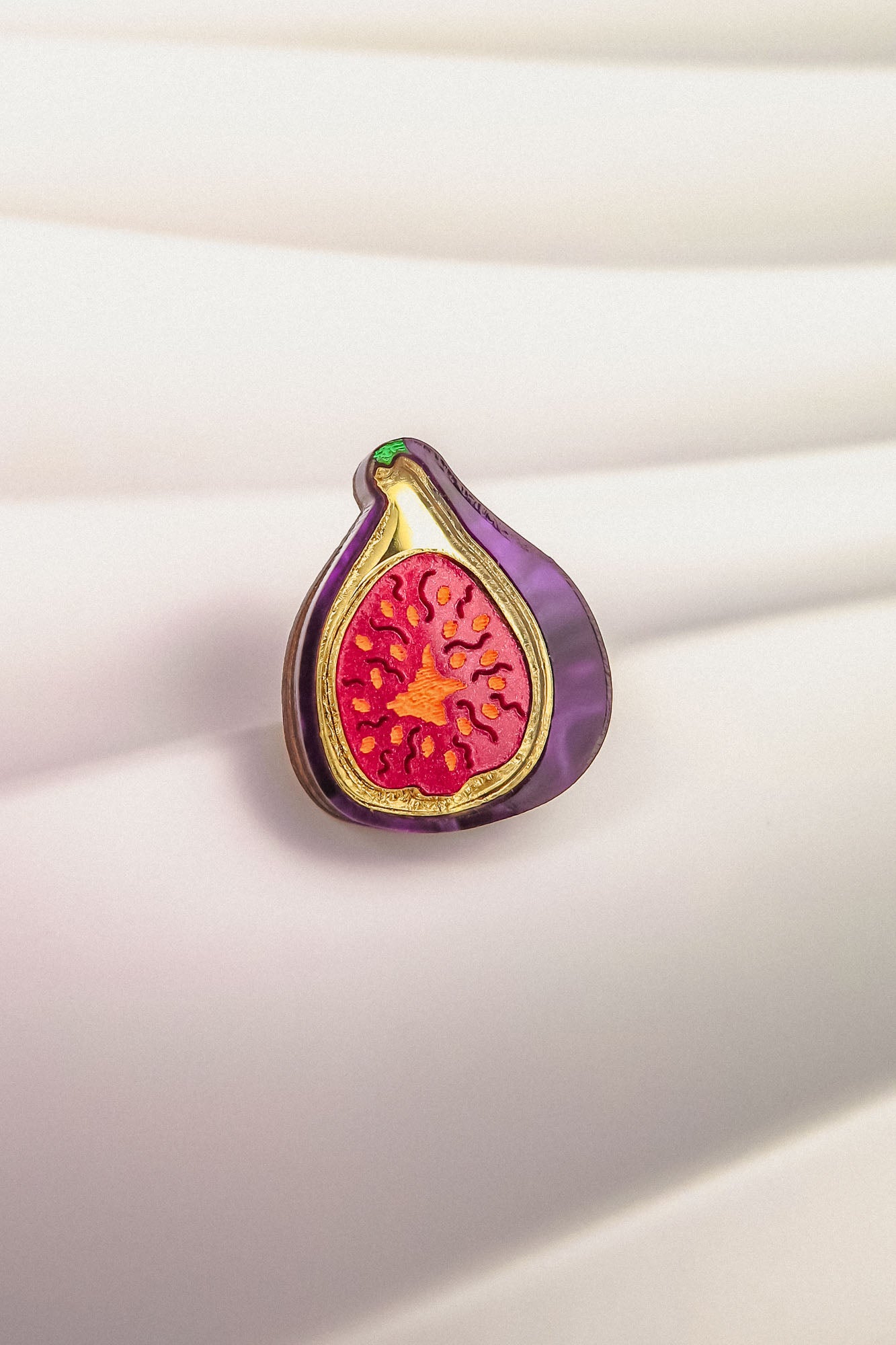 A colorful Fig Pin shaped like a fig, with a purple outer edge and a vibrant red and yellow interior, is displayed on a softly lit, white, textured background—a handcrafted, lightweight accessory perfect for any collection.