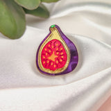 A handcrafted Fig Pin shaped like a sliced fig, featuring a purple border and red center, rests on silky white fabric, with green leaves in the upper left corner—a unique and lightweight accessory for your collection.