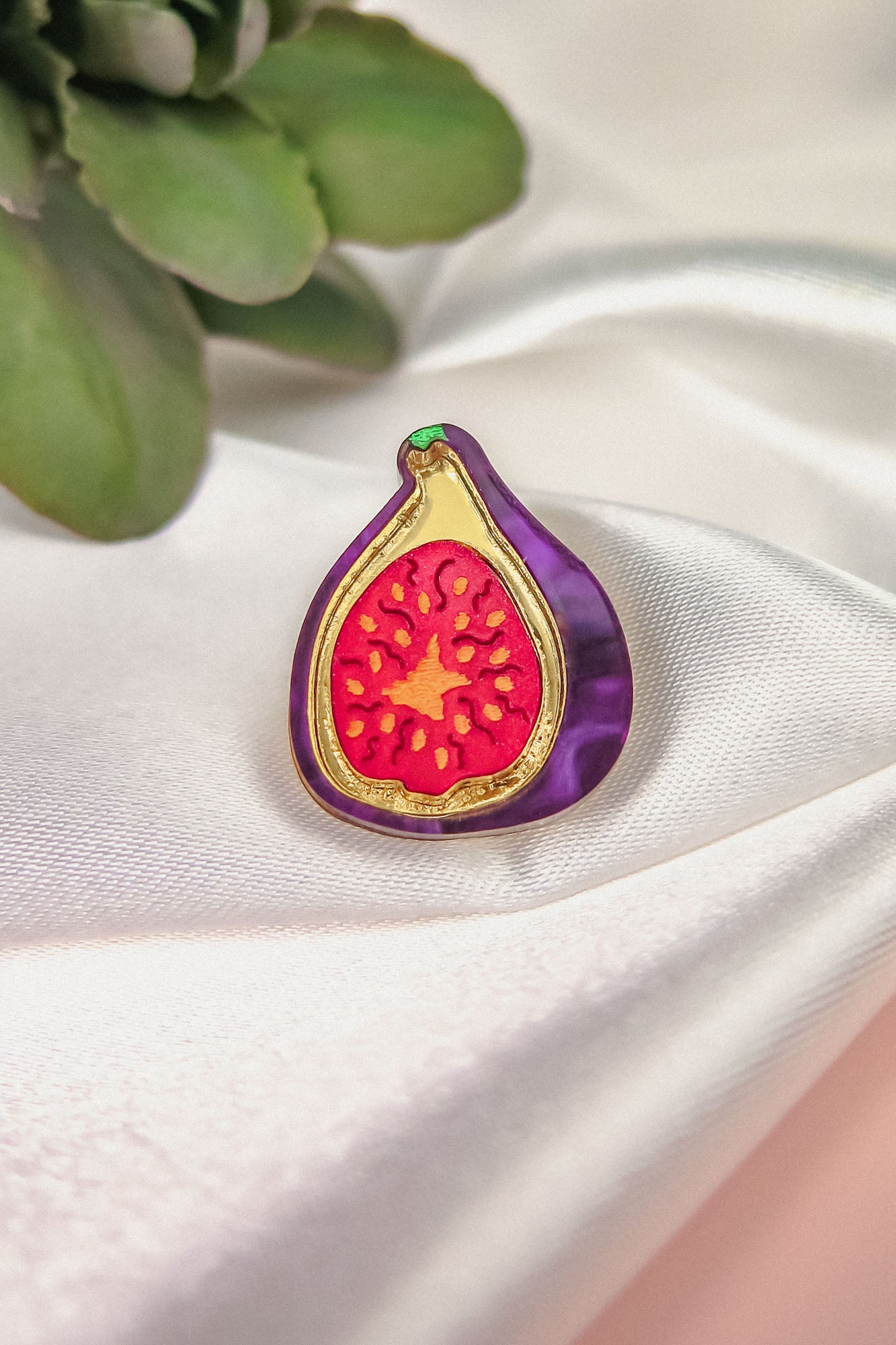 A handcrafted Fig Pin shaped like a sliced fig, featuring a purple border and red center, rests on silky white fabric, with green leaves in the upper left corner—a unique and lightweight accessory for your collection.