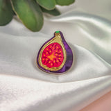 A handcrafted Fig Pin, this colorful enamel pin features a purple edge and vibrant red interior, displayed on white satin with green leaves behind. A lightweight accessory that adds a pop of color to any look.