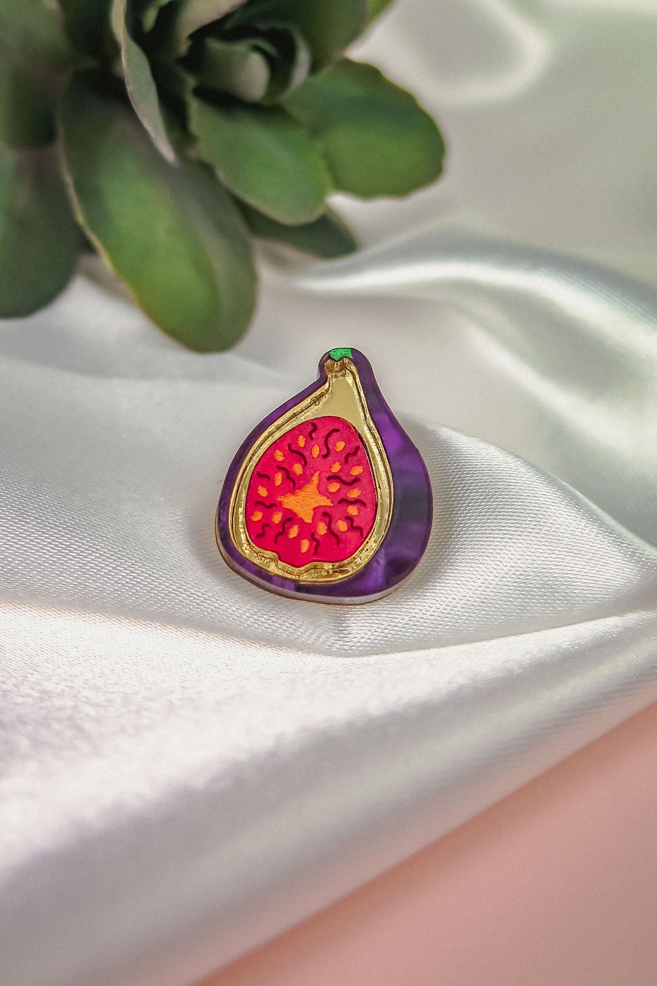 A handcrafted Fig Pin, this colorful enamel pin features a purple edge and vibrant red interior, displayed on white satin with green leaves behind. A lightweight accessory that adds a pop of color to any look.